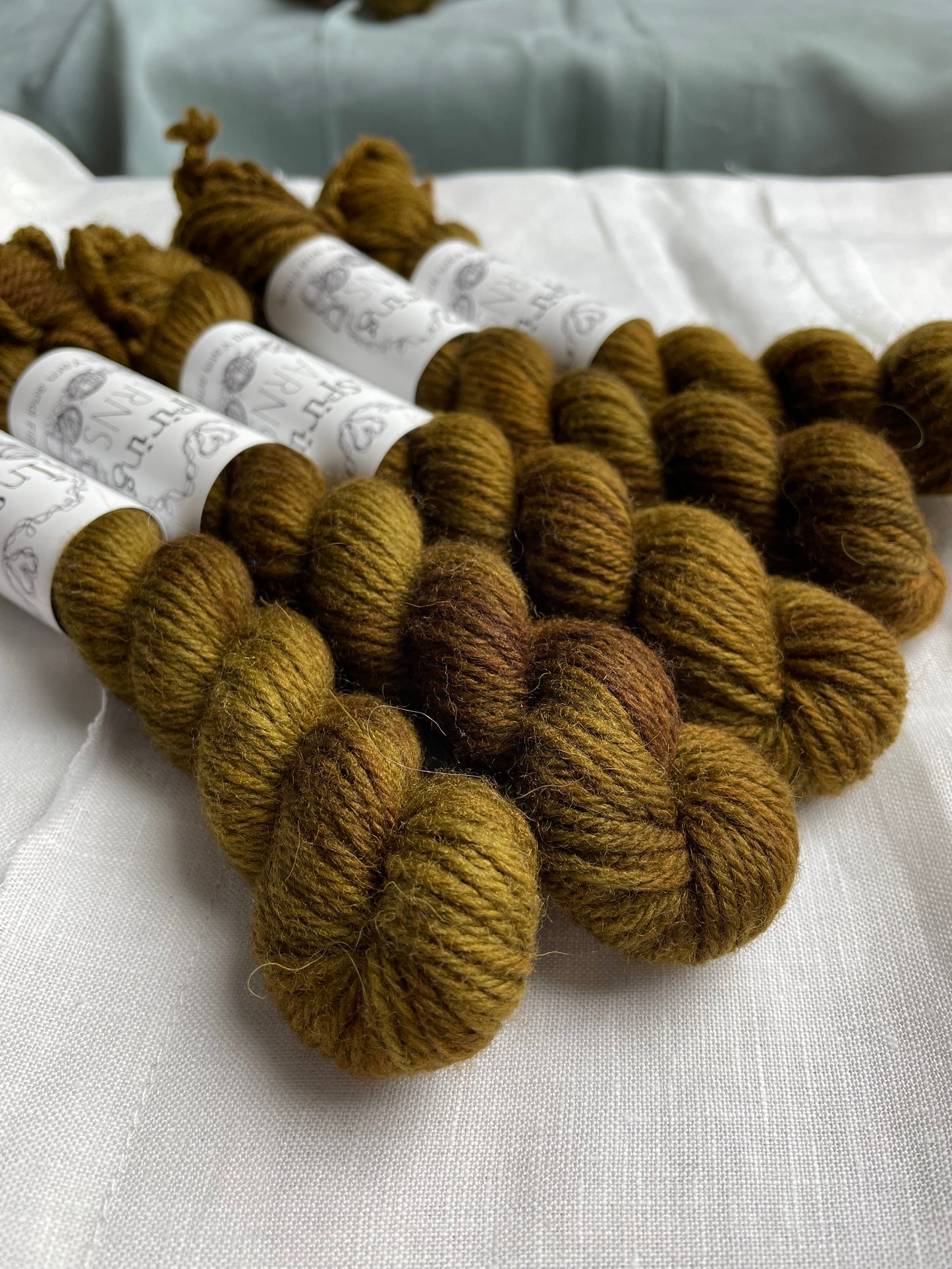 Close up of five mini skeins of a tonal green brown Highland DK yarn branded Inspiring Yarns on a white linen background.