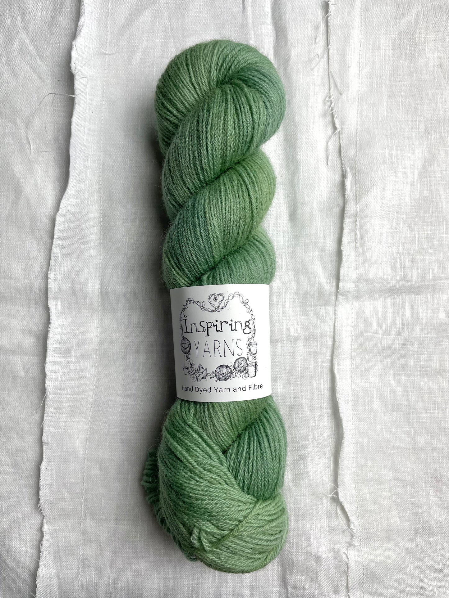 A skein of soft mint green coloured Highland 4Ply yarn branded Inspiring Yarns on a white linen background.