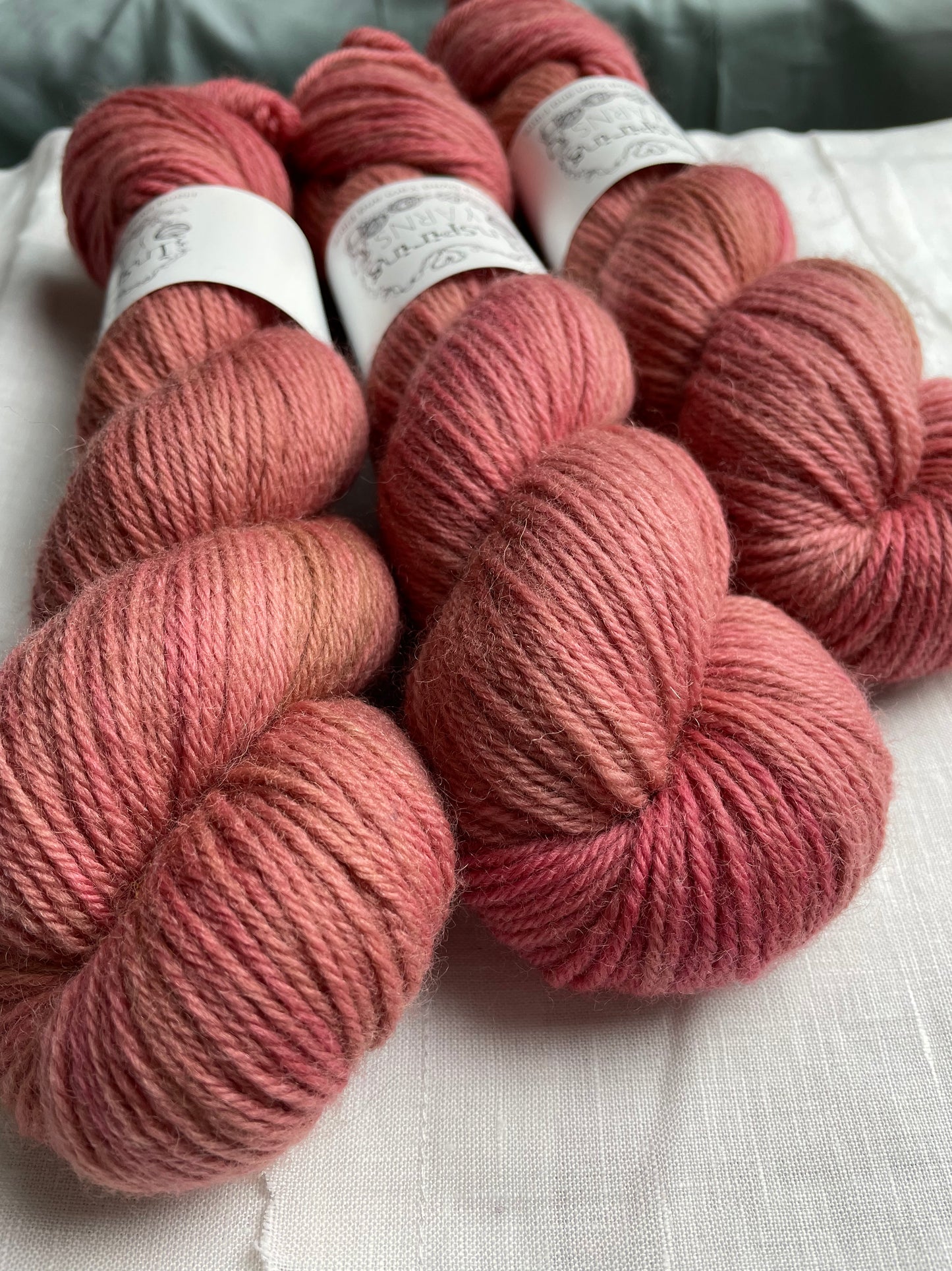 Close up of three skeins of a tonal dusky pink Highland DK yarn branded Inspiring Yarns on a white linen background.