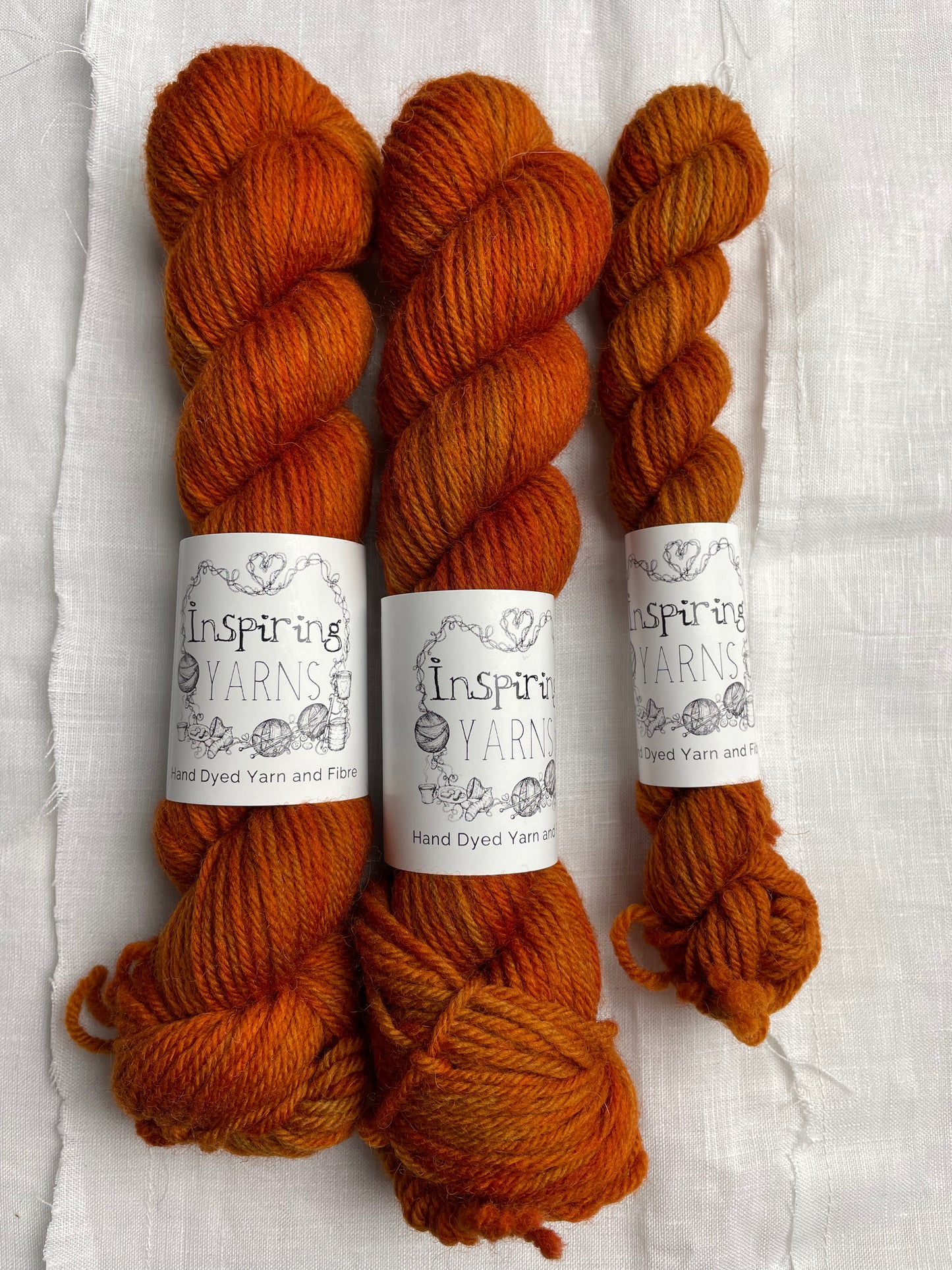 Three skeins of a tonal burnt orange Highland DK yarn branded Inspiring Yarns on a white linen background.