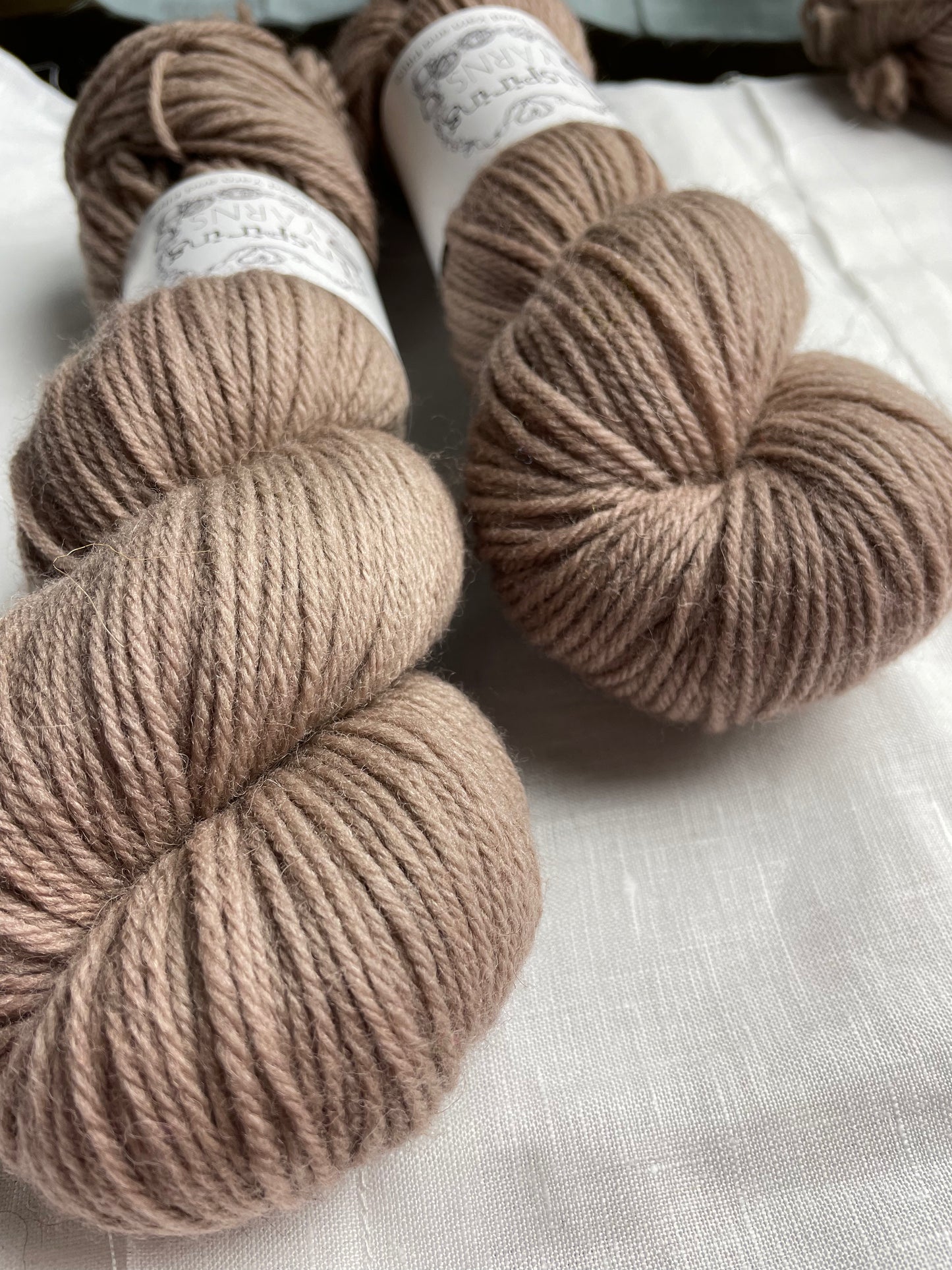 Close up of two skeins of tonal taupe coloured Highland DK yarn branded Inspiring Yarns on a white linen background.
