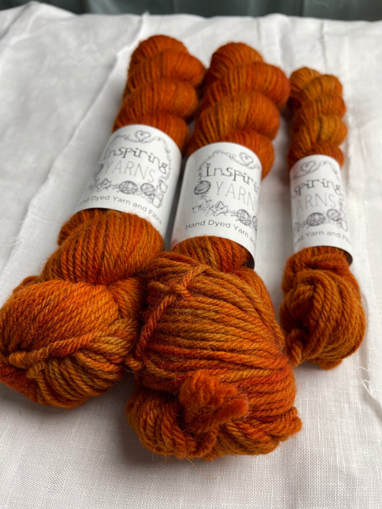 Close up of three skeins of a tonal burnt orange Highland DK yarn branded Inspiring Yarns on a white linen background.