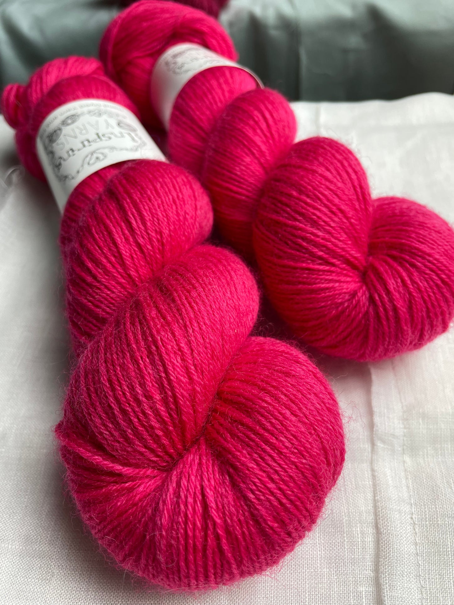 Close up of two skeins of tonal bright cerise pink coloured Highland 4Ply yarn branded Inspiring Yarns on a white linen background.