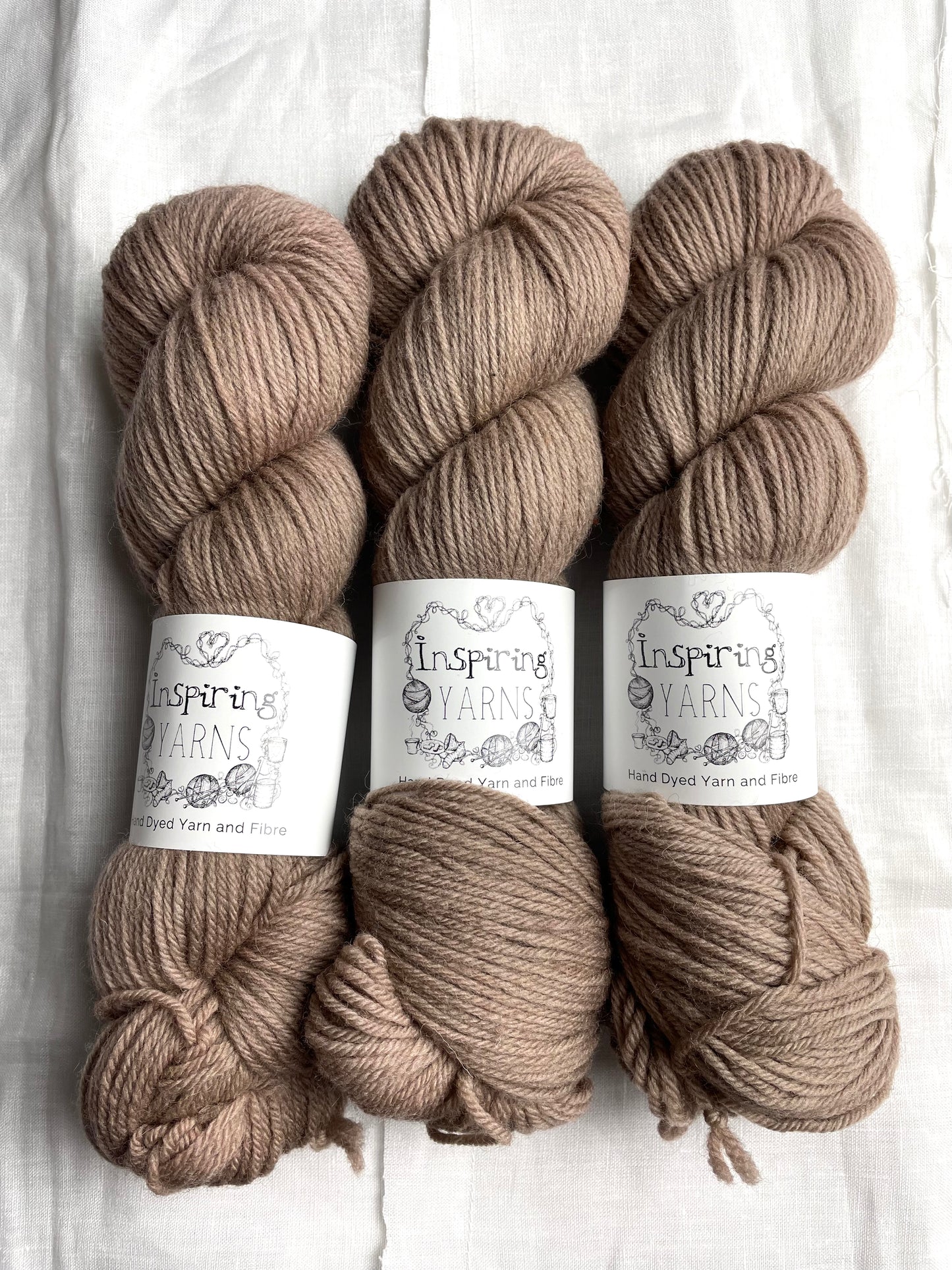 Three skeins of tonal taupe coloured Highland DK yarn branded Inspiring Yarns on a white linen background.