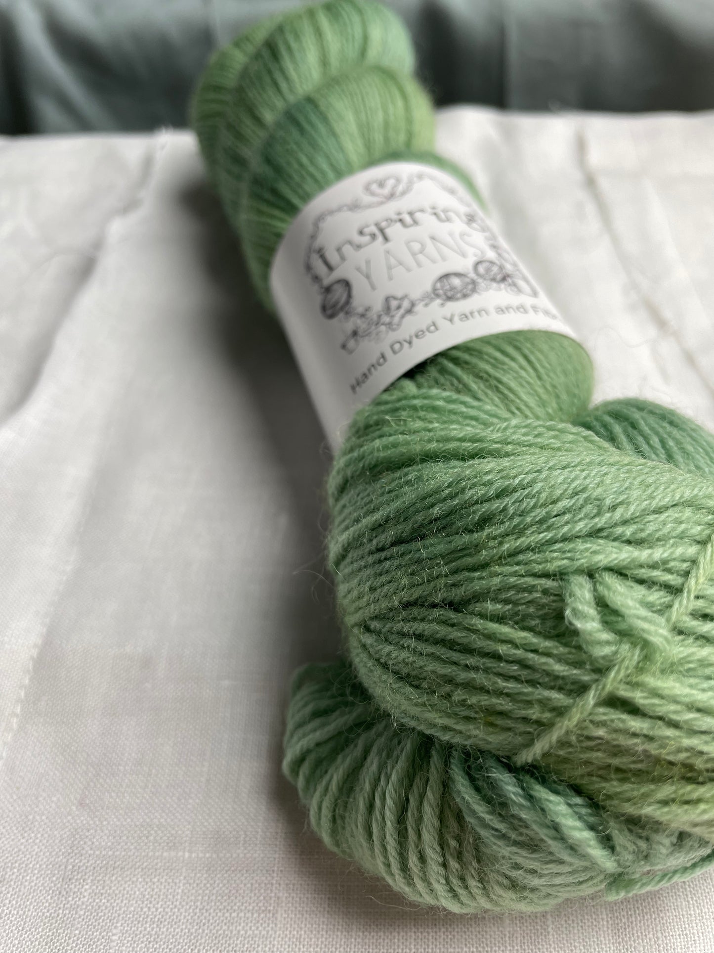 Close up of a skein of soft mint green coloured Highland 4Ply yarn branded Inspiring Yarns on a white linen background.