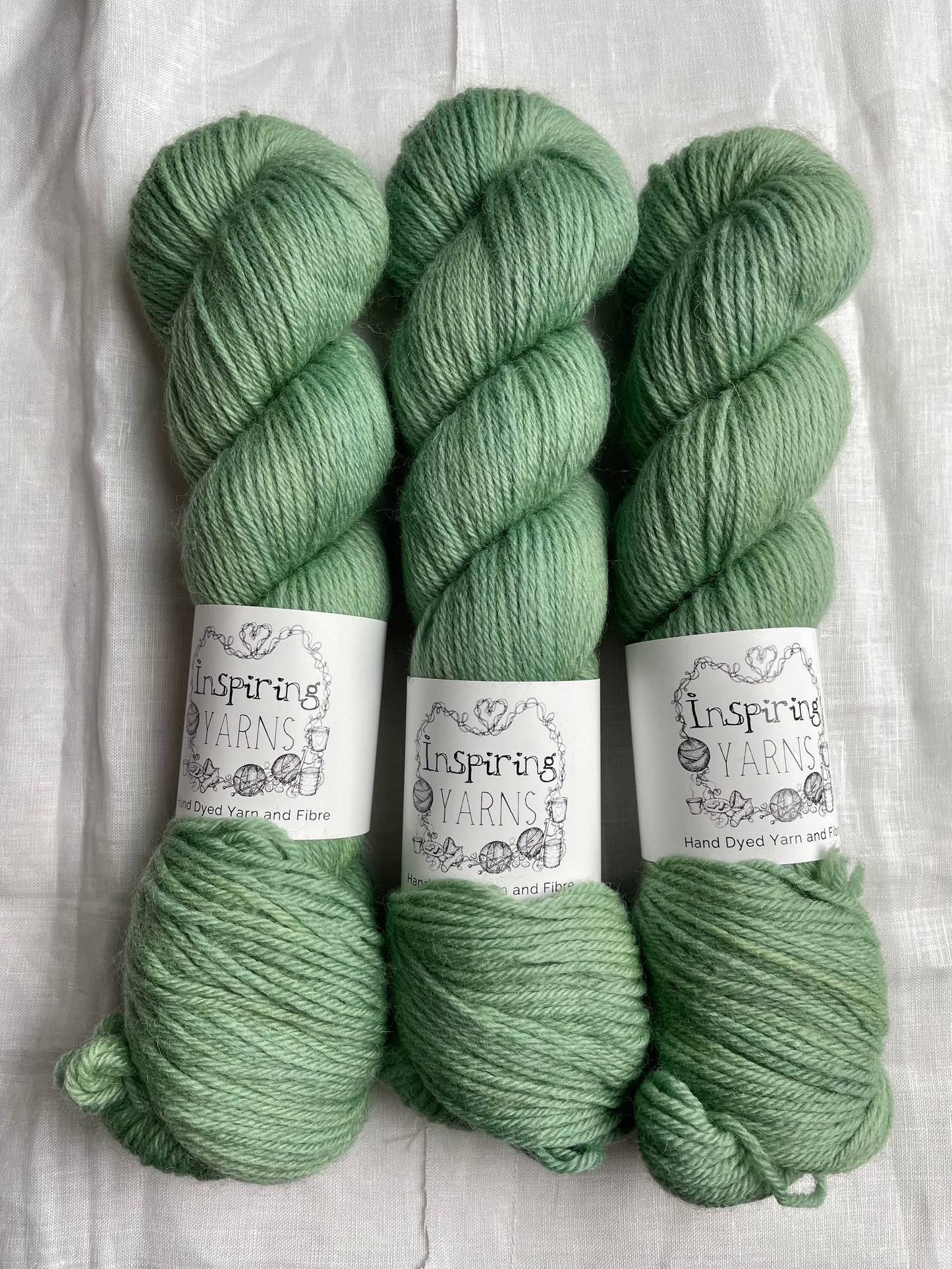 Three skeins of soft mint green coloured Highland DK yarn branded Inspiring Yarns on a white linen background.