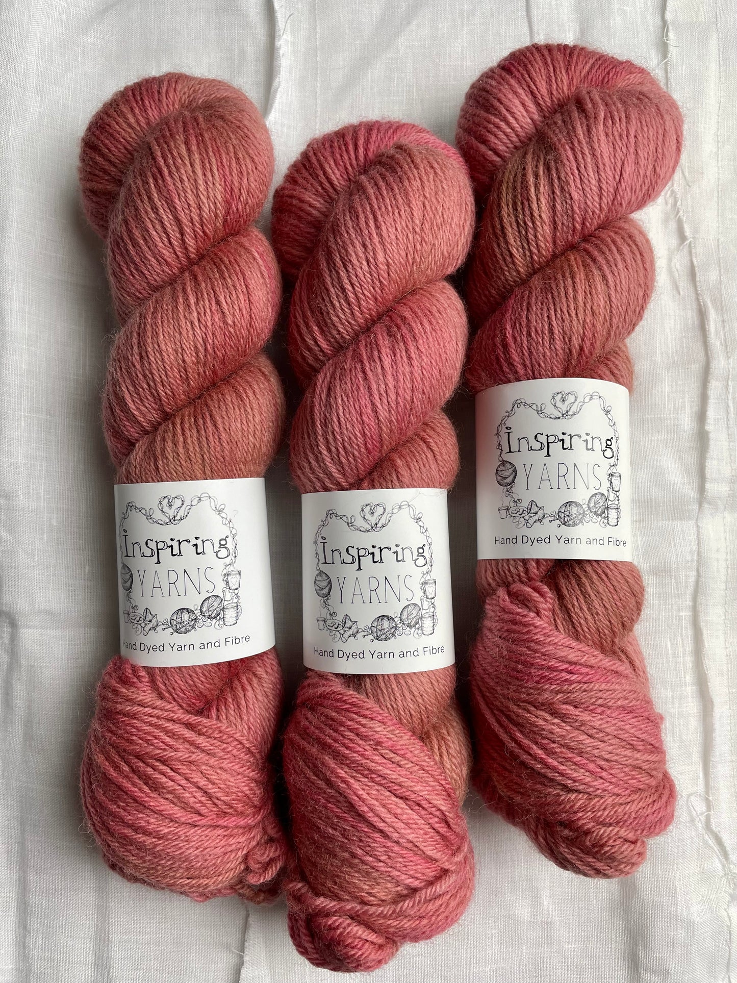 Three skeins of a tonal dusky pink Highland DK yarn branded Inspiring Yarns on a white linen background.