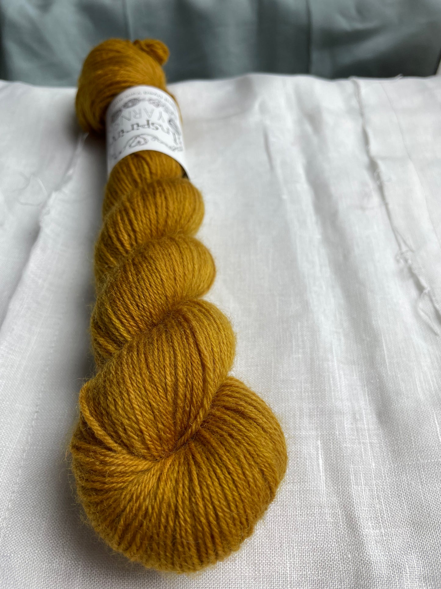Close up of a skein of tonal mustard yellow coloured Highland 4Ply yarn branded Inspiring Yarns on a white linen background.
