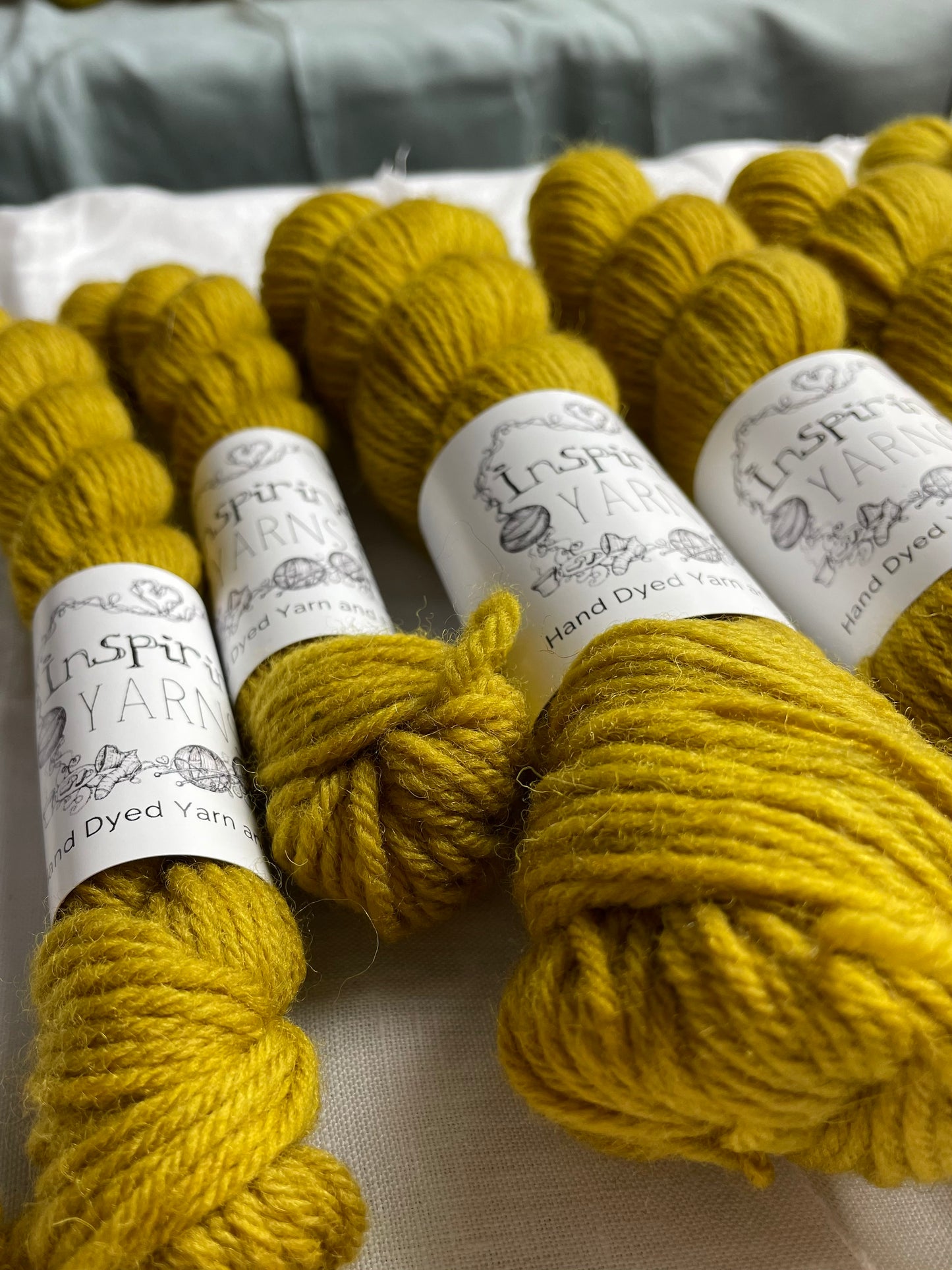 Close up of skeins of mixed sizes of tonal golden ochre coloured Highland DK yarn branded Inspiring Yarns on a white linen background.