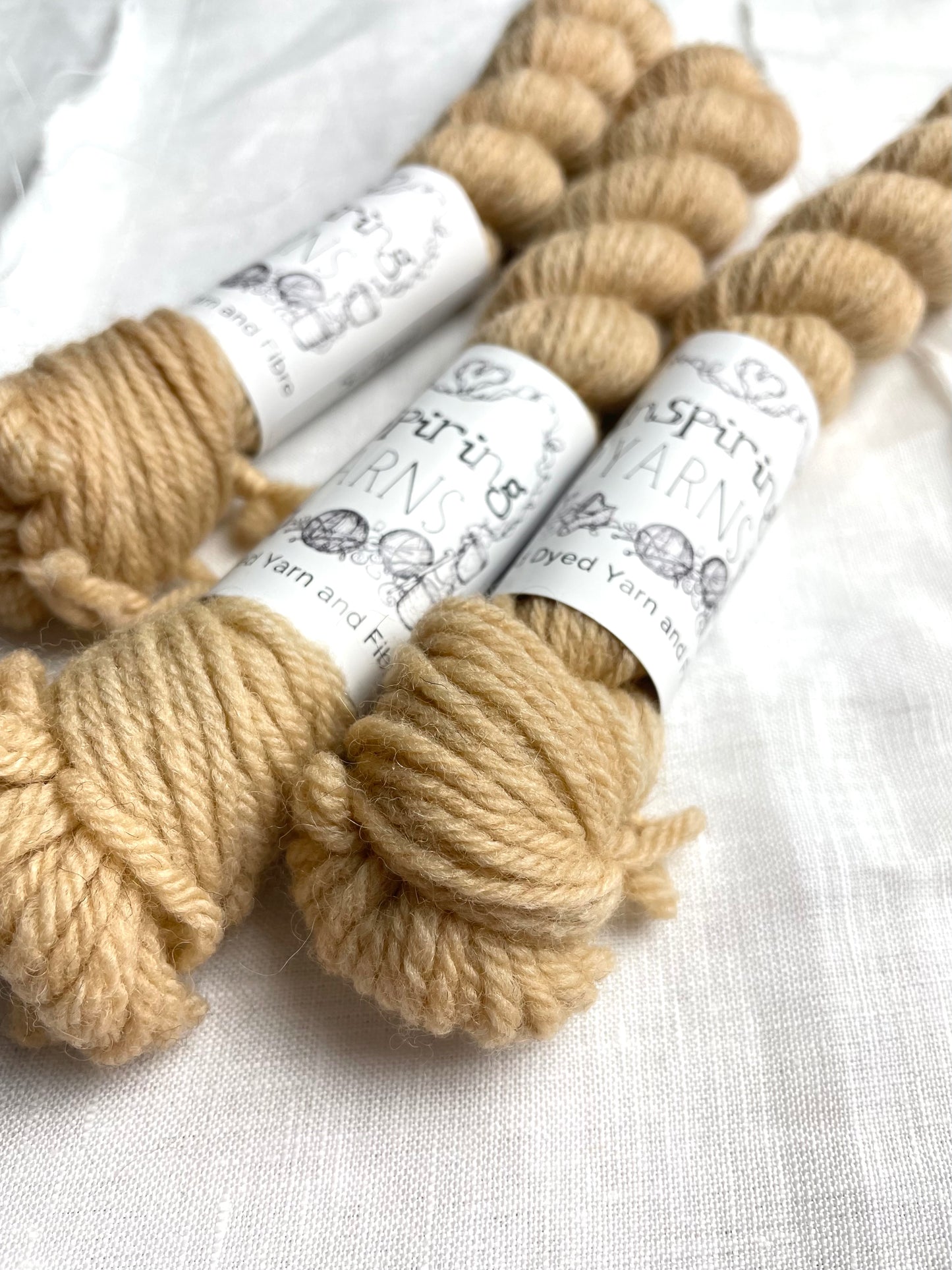 Close up of three mini skeins of soft golden cream coloured Highland DK yarn branded Inspiring Yarns on a white linen background.