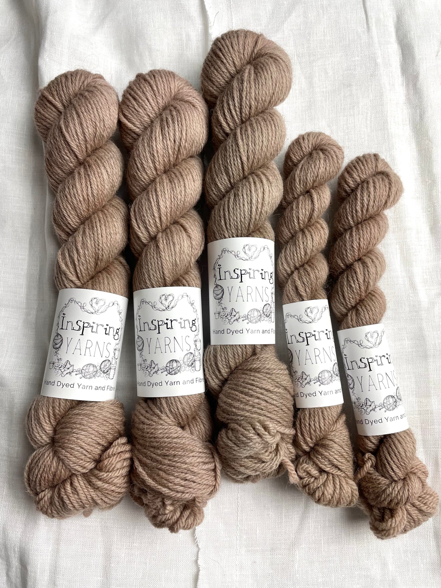 Skeins of mixed sizes of tonal taupe coloured Highland DK yarn branded Inspiring Yarns on a white linen background.