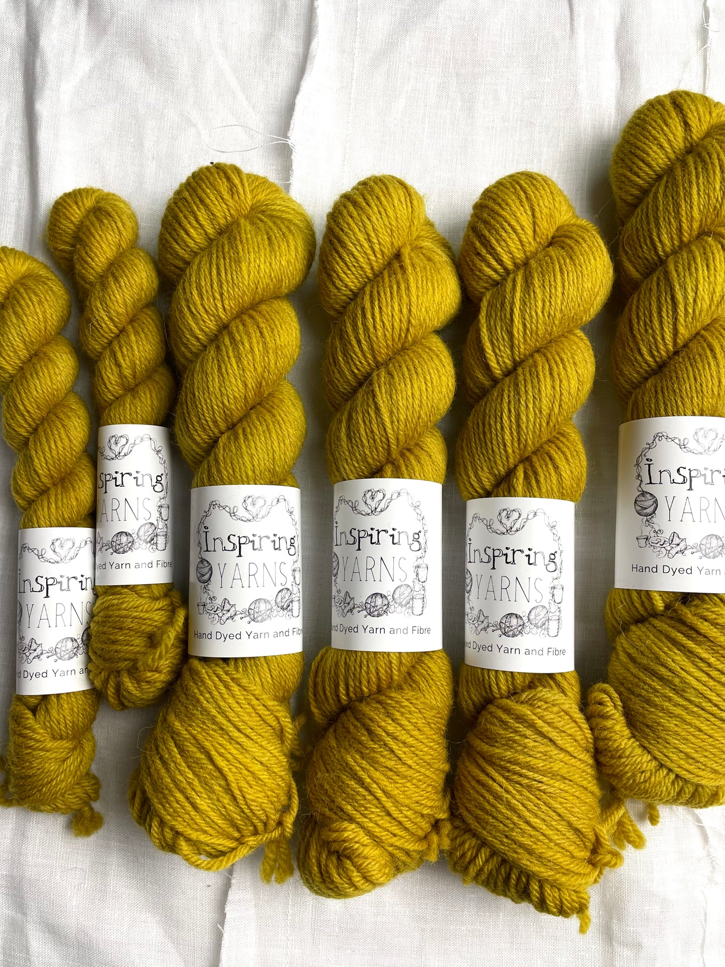 Skeins of mixed sizes of tonal golden ochre coloured Highland DK yarn branded Inspiring Yarns on a white linen background.