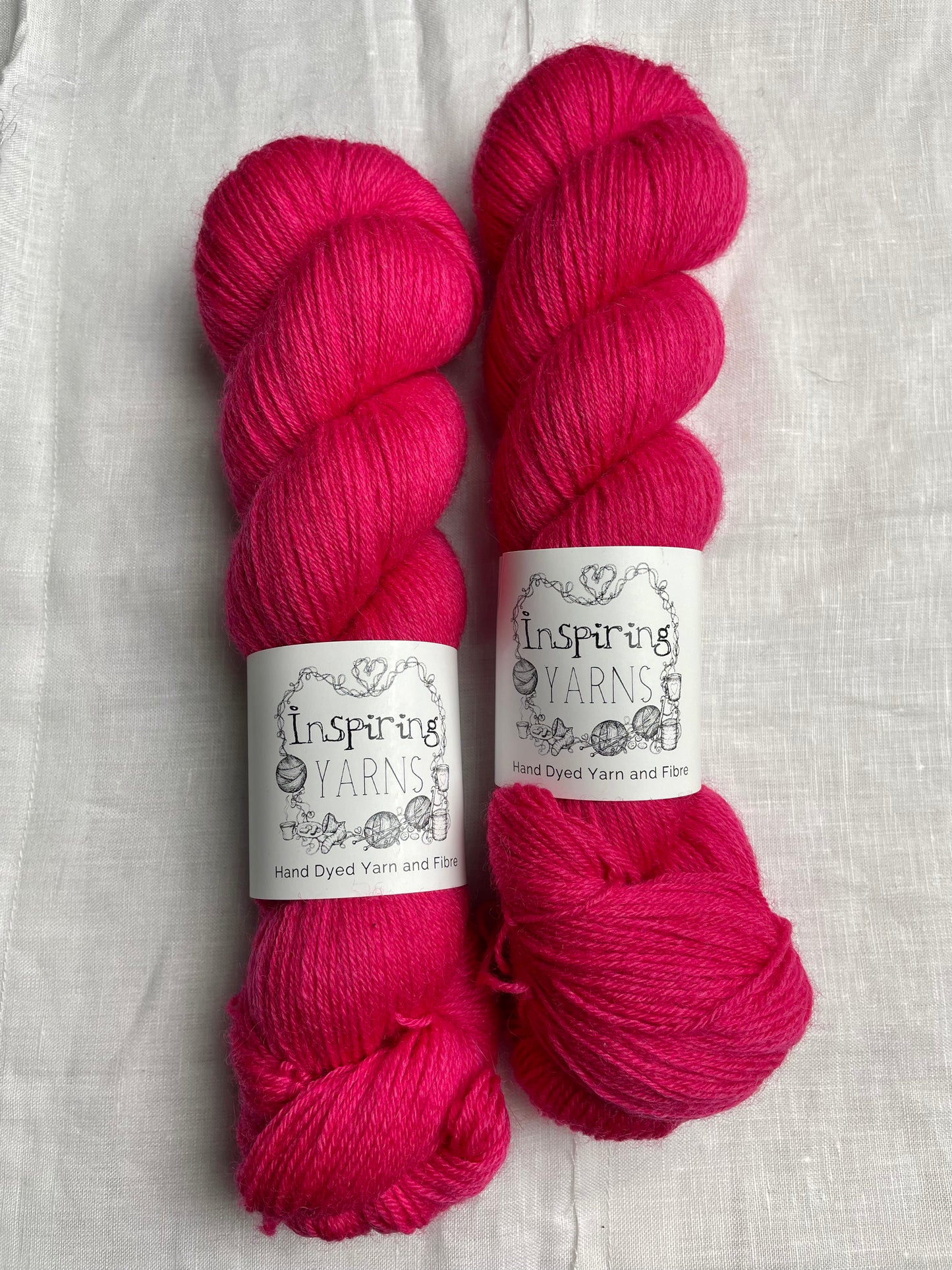 Two skeins of tonal bright cerise pink coloured Highland 4Ply yarn branded Inspiring Yarns on a white linen background.