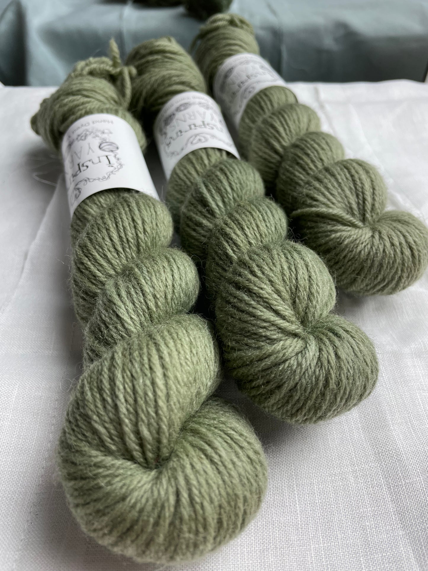 Close up of three skeins of a tonal light green grey Highland DK yarn branded Inspiring Yarns on a white linen background.