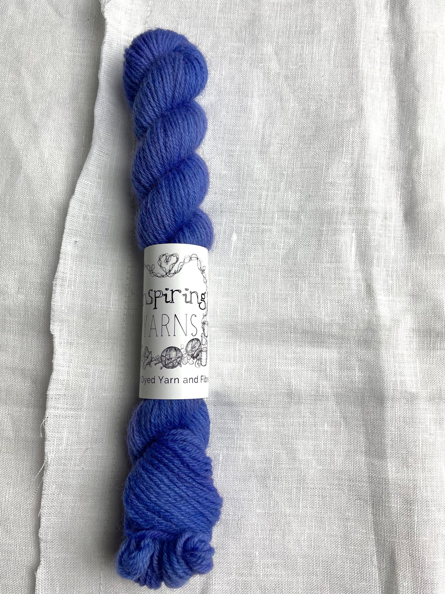 A mini skein of tonal bright blue lilac coloured Highland 4Ply yarn branded Inspiring Yarns on a white linen background.