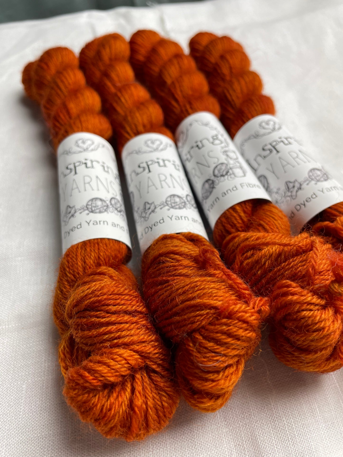 Four mini skeins of tonal burnt orange coloured Highland 4Ply yarn branded Inspiring Yarns on a white linen background.