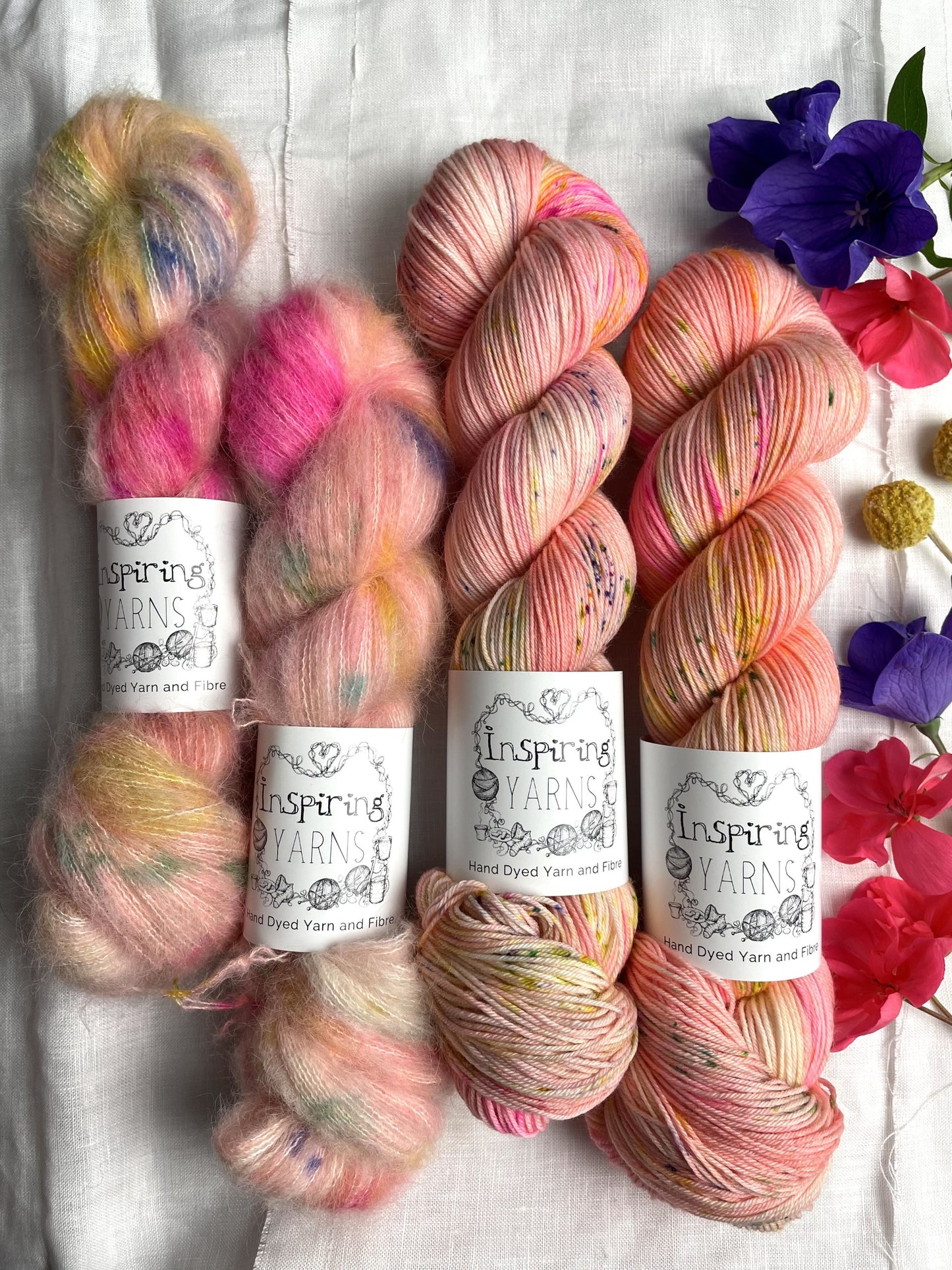 Four skeins of yarn in a soft peach speckled colourway on a white linen background with some real fowers. The two skeins on the left are Brushed Suri Alpaca Lace and the two on the right are Merino Smooth Sock.