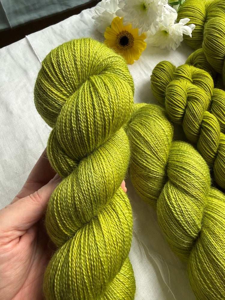 A hand holding a skein of bright green BFL Silk Lace yarn, in the background some other yarn skeins lie on a white background with some flowers. 