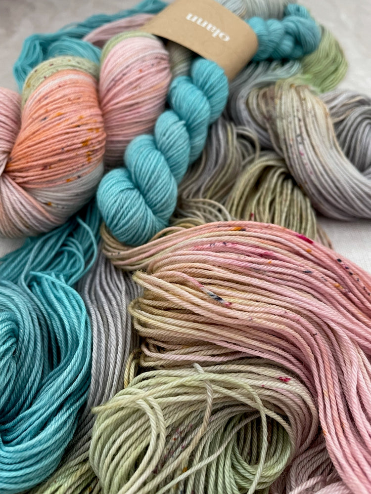 a skein of multi coloured yarn in pastel shades lies loose on a table, a twisted skein of the same yarn lies on top with a light turquoise mini, the yarn band reads Olann 