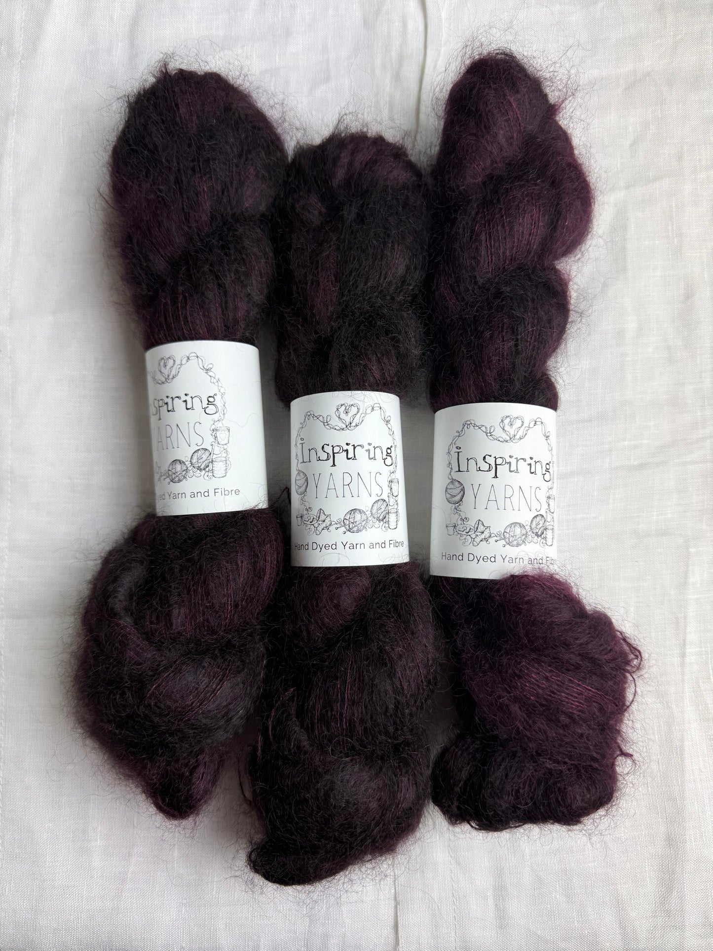 Three skeins of dark burgundy mohair yarn labelled Inspiring Yarns on a white linen fabric background