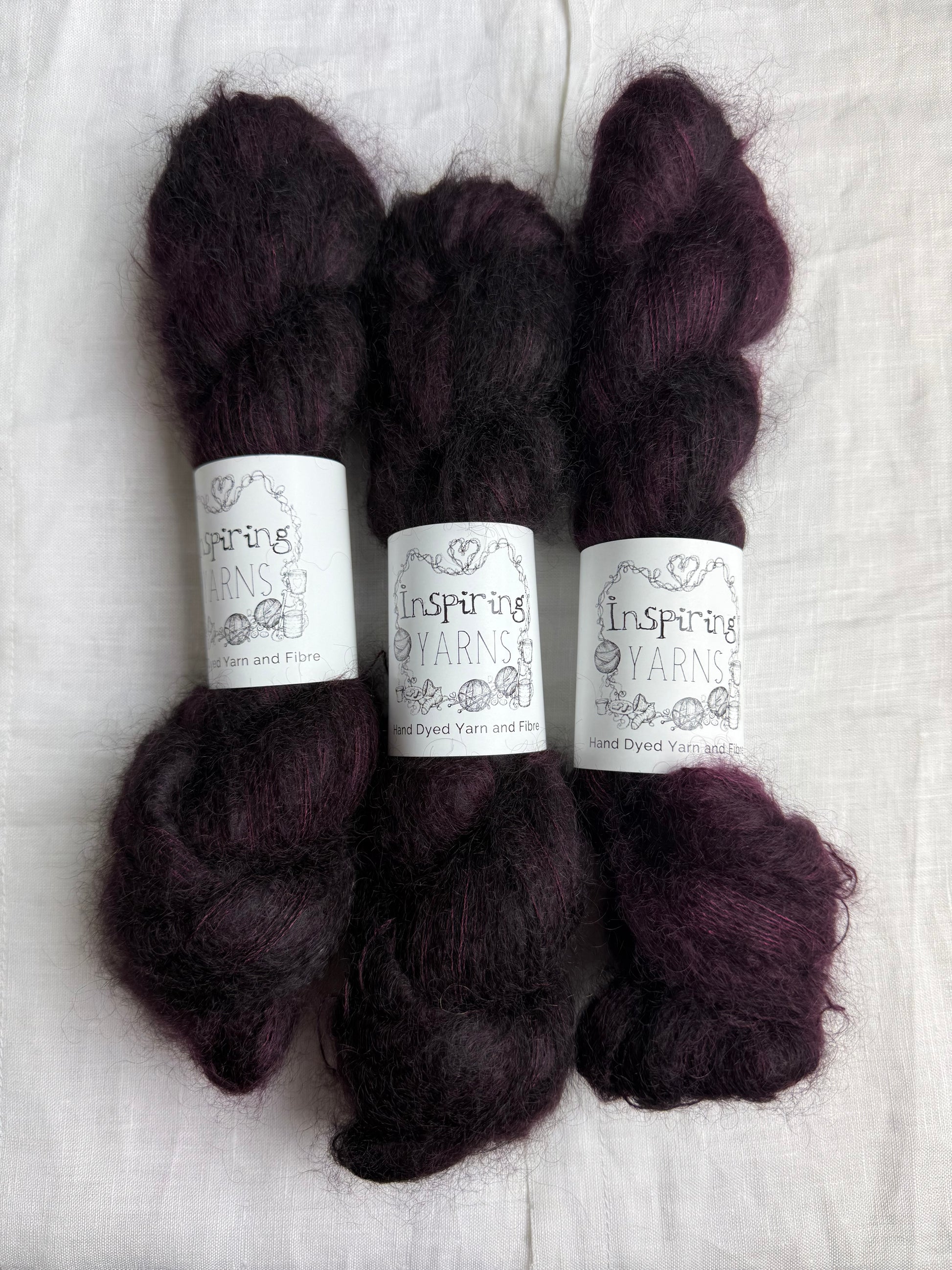 Three skeins of dark burgundy mohair yarn labelled Inspiring Yarns on a white linen fabric background