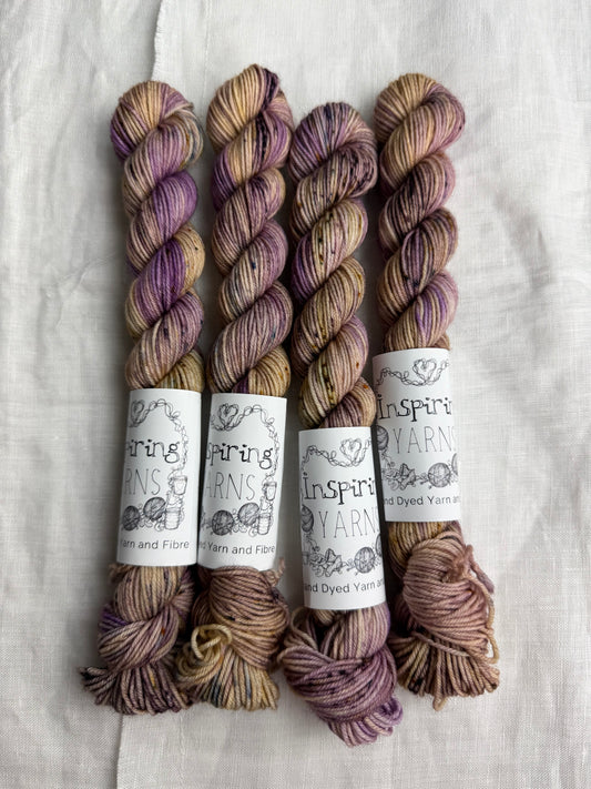 Four skeins of cream and lilac with speckles in deep purple and earthy browns with Inspiring Yarns labels on a light textured background.
