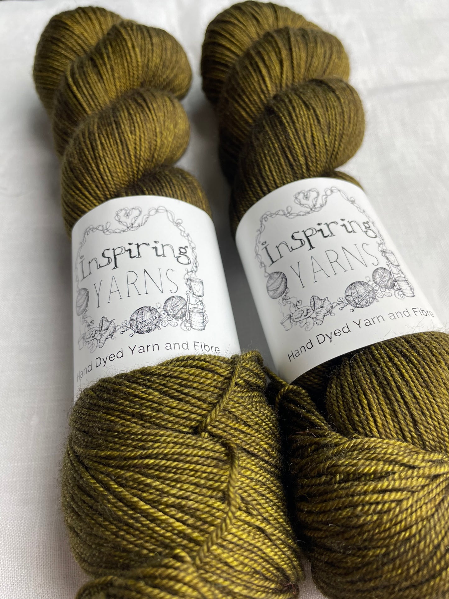 Close up of two tonal dark olive green coloured skeins of hand dyed Yak Silk 4Ply yarn branded Inspiring Yarns on a white linen background.