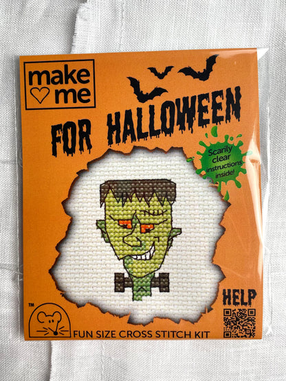 A mini crosstitch kit of a face in the style of Frankenstein’s monster photographed on a white linen background.  It is branded Mouseloft Make Me For Halloween in orange packaging.