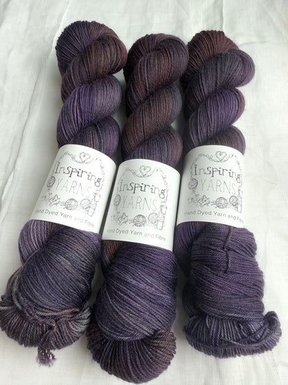 three skeins of dark purple yarn with orange and blue tones with inspiring yarns label and white background.