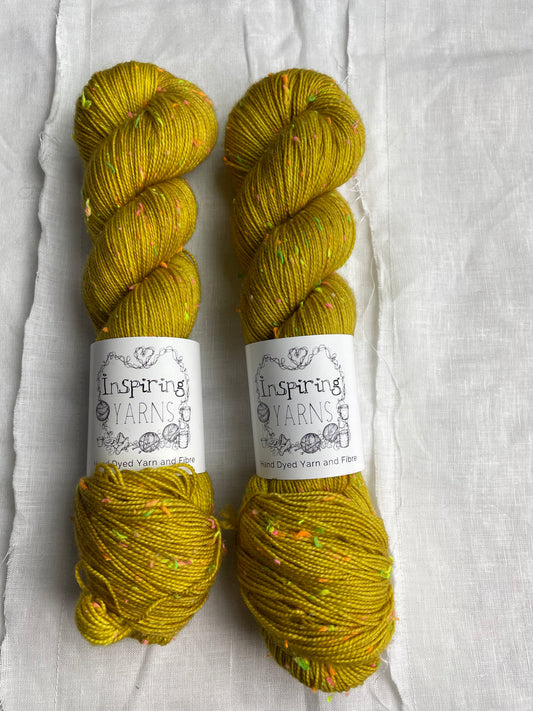 Two skeins of tonal golden ochre coloured hand dyed Disco Donegal yarn branded Inspiring Yarns on a white linen background.  The yarn has multi coloured neon neps.