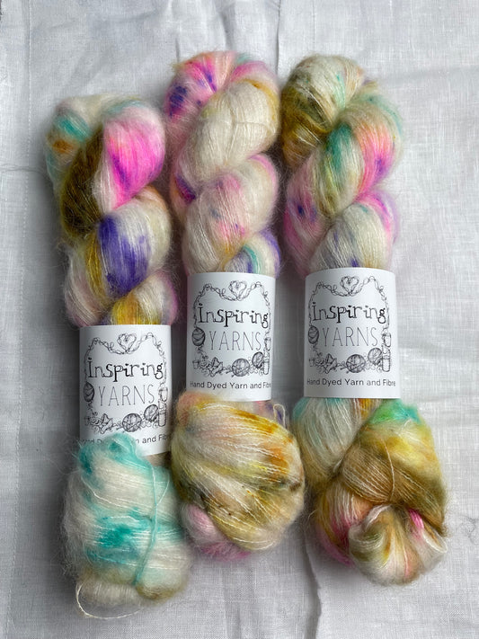 Three skeins of brightly speckled natural coloured hand dyed fluffy Alpaca Silk Lace yarn labelled Inspiring Yarns on a white linen background. 