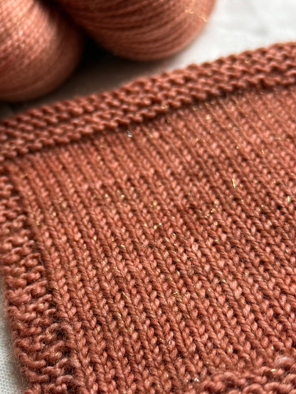 Close-up of a knitted fabric swatch in tonal warm terracotta yarn, with matching yarn balls blurred in the background.