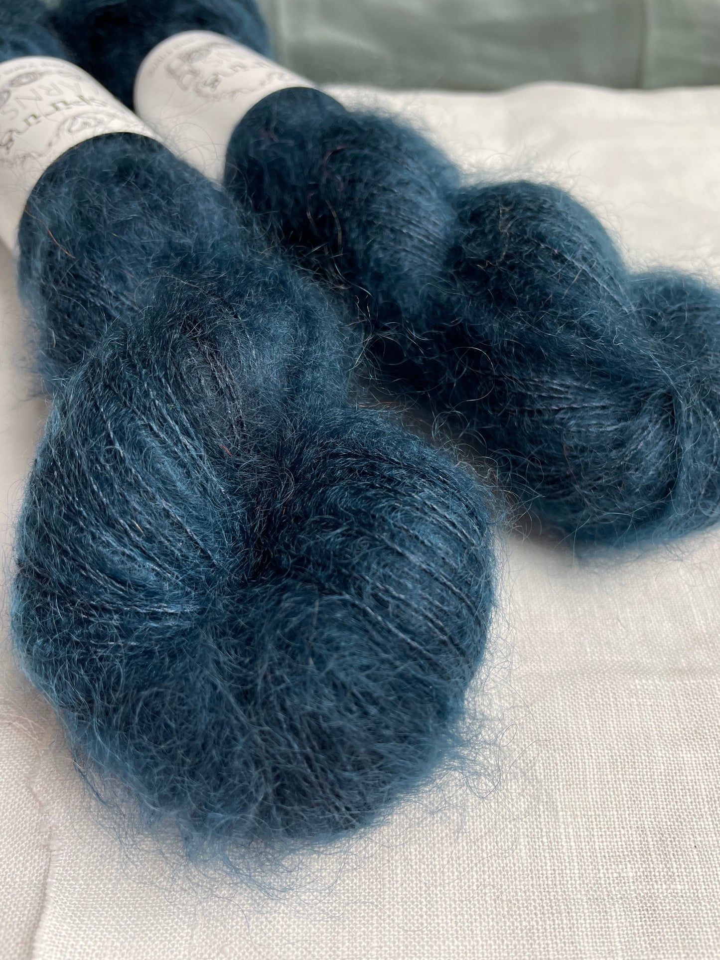 Close up of 2 skeins of dark steel blue mohair silk lace yarn labelled Inspiring Yarns on a white linen background.