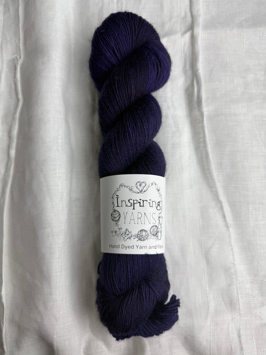 a skein of dark purple/blue yarn with black and white inspiring yarns label around it on a plain white fabric background.