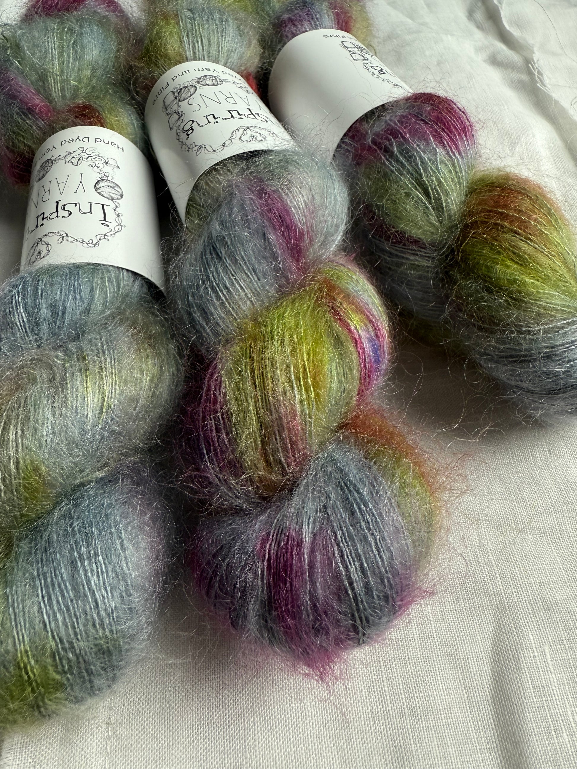 Three skeins of yarn with blueish grey tones with reds greens and browns speckled through labels on a white background.