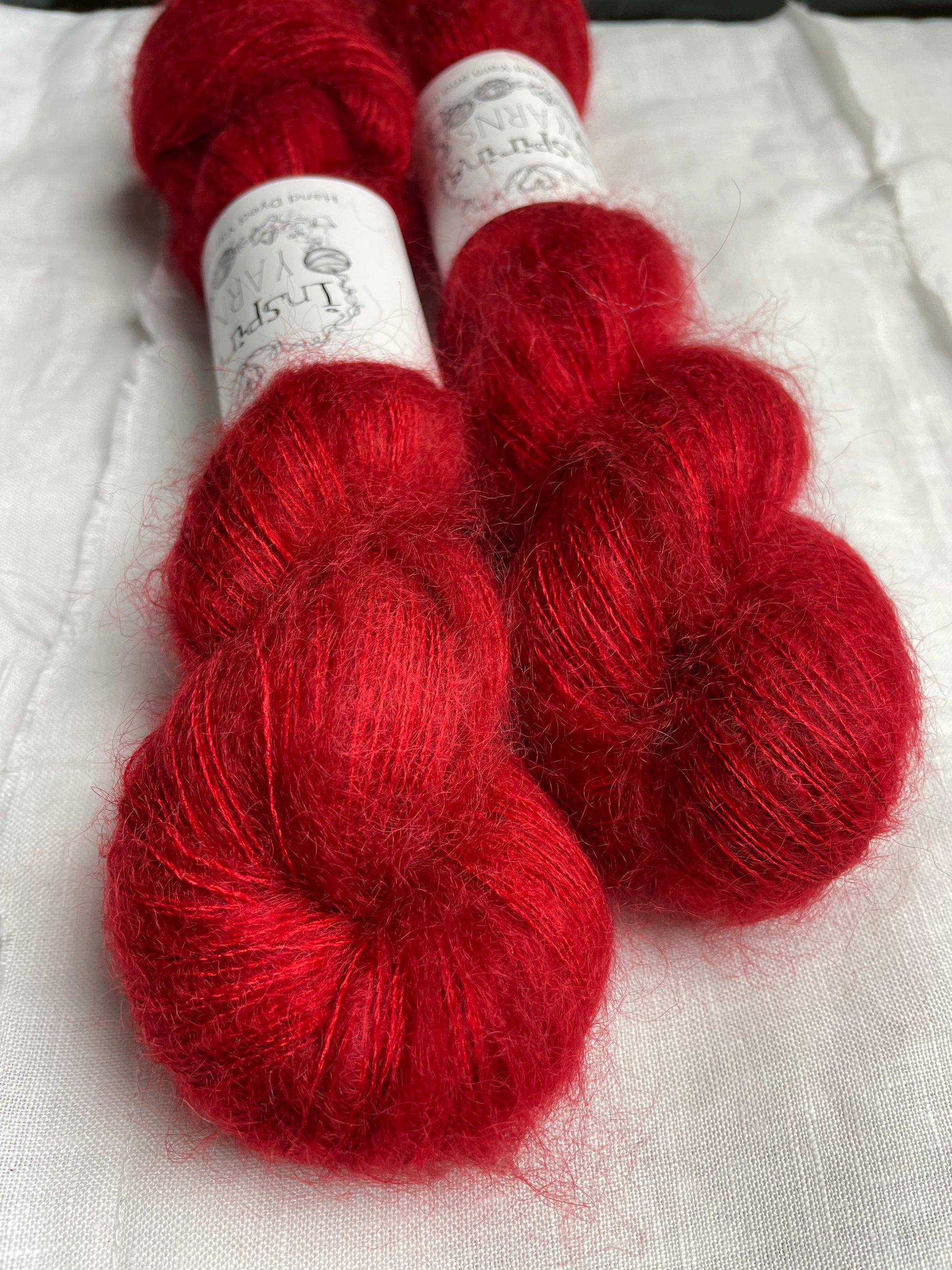 Closer up of two skeins of tonal bright red kid mohair silk lace yarn labelled Inspiring Yarns on a white linen background.