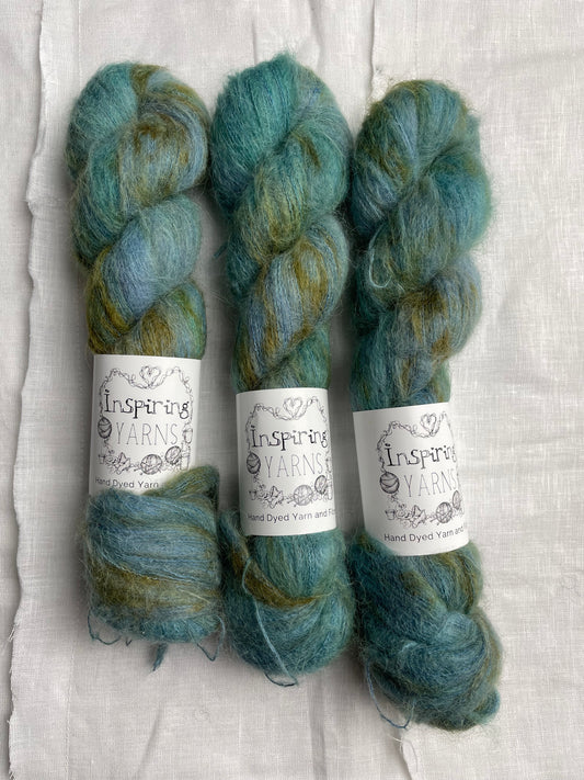 Three skeins of speckled deep teal coloured hand dyed fluffy Alpaca Silk Lace yarn labelled Inspiring Yarns on a white linen background. 