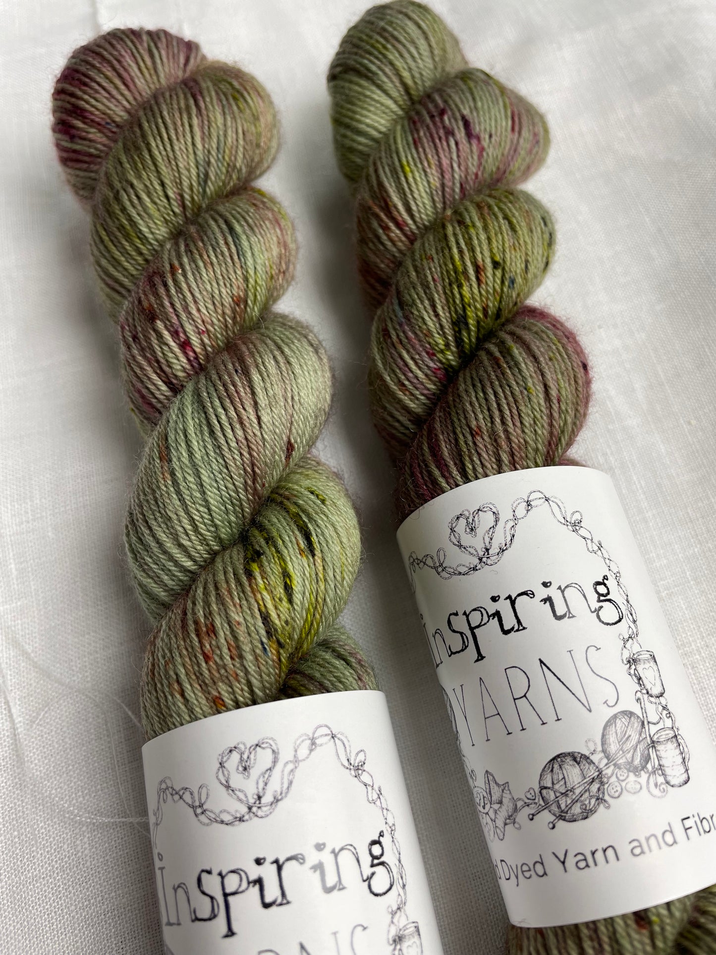 Two skeins of Cassidy, a sage green with speckles coloured Merino Smooth Sock, labelled Inspiring Yarns on a white linen background. 