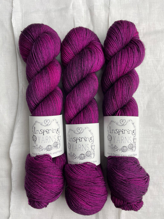 Three lightly variegated cerise pink and dark purple coloured skeins of Merino Smooth Sock yarn branded  Inspiring Yarns on a white linen fabric background. 