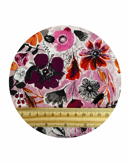 Round fabric swatch of Night Bloom Rose from the Eerie Collection by Art Gallery Fabrics, a floral pattern with colourful flowers and leaves on a light background in pinks, black, grey and orange, with a wooden ruler in centrimetres for scale..