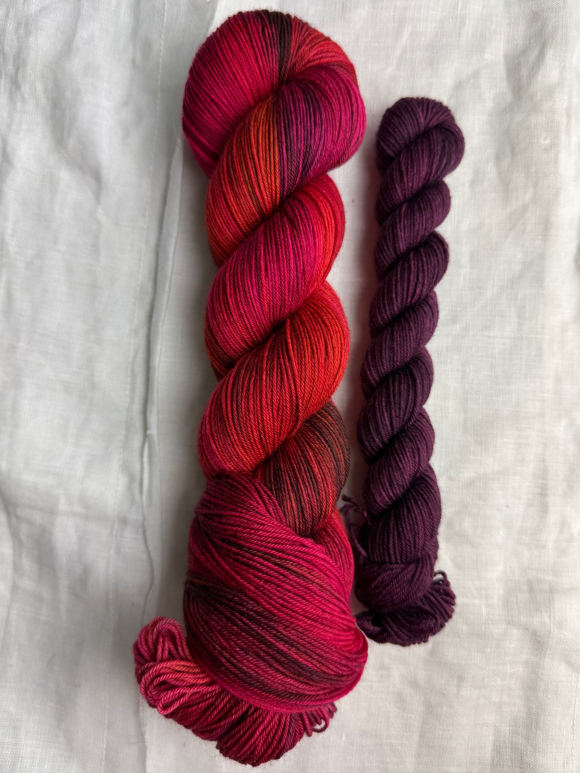 Variegated red, pink, orange and purple yarn skein with a burgundy mini skeinon a white fabric background