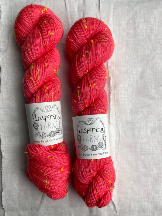 Two skeins of tonal dark neon coral coloured hand dyed Disco Donegal yarn branded Inspiring Yarns on a white linen background.  The yarn has multi coloured neon neps.