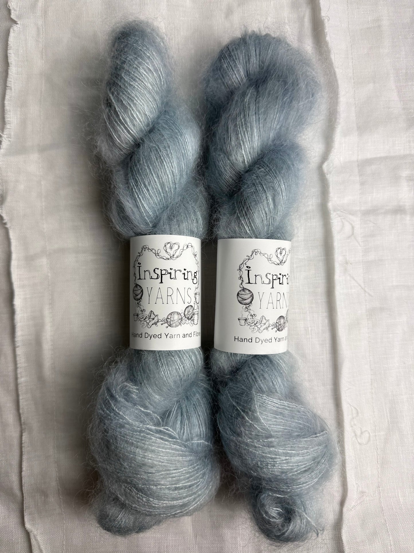 Two skeins of gray yarn with 'Inspiring Yarns' labels on a textured fabric background.