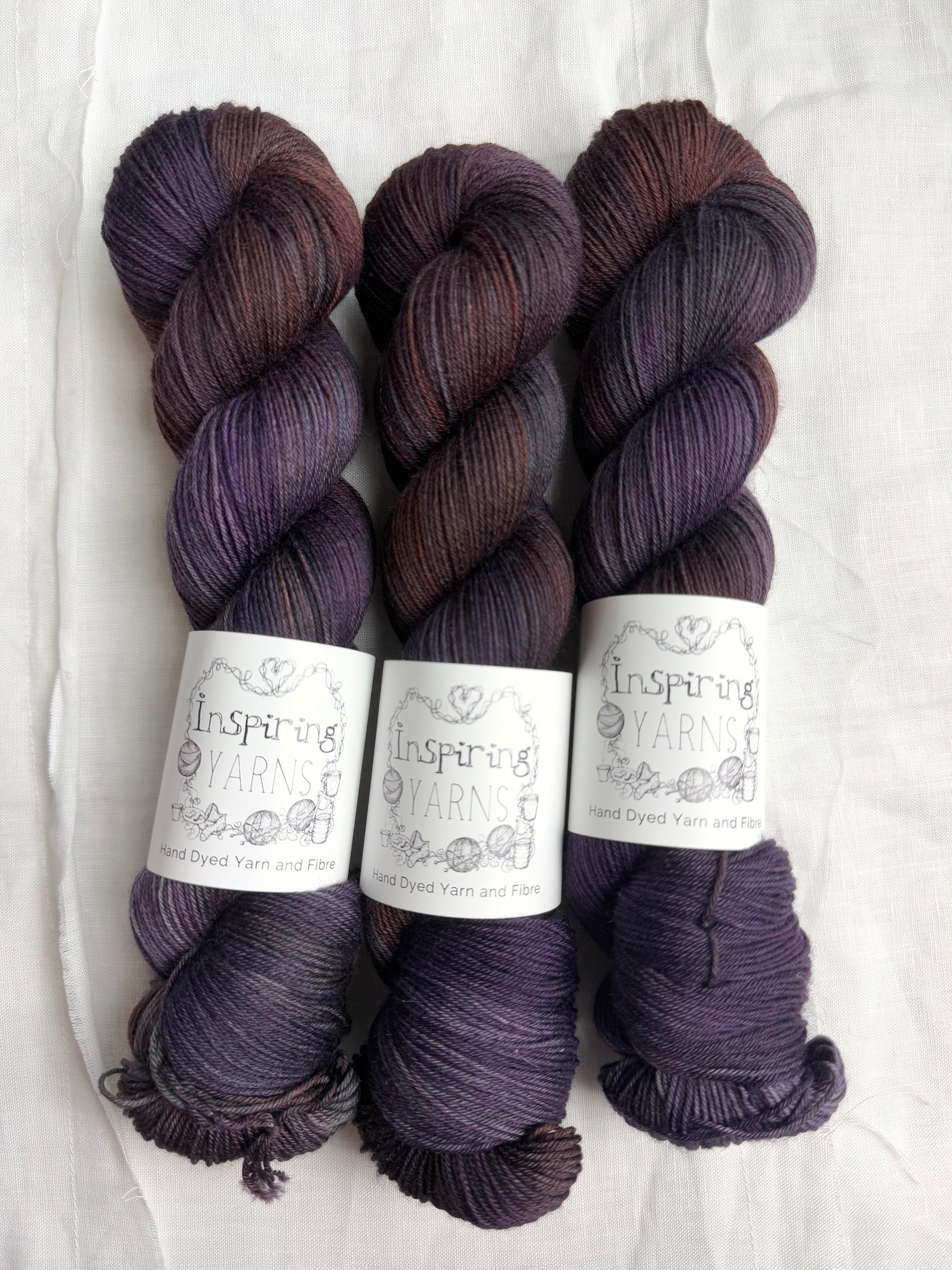 three skeins of dark purple yarn with orange and blue tones with inspiring yarns label and white background.