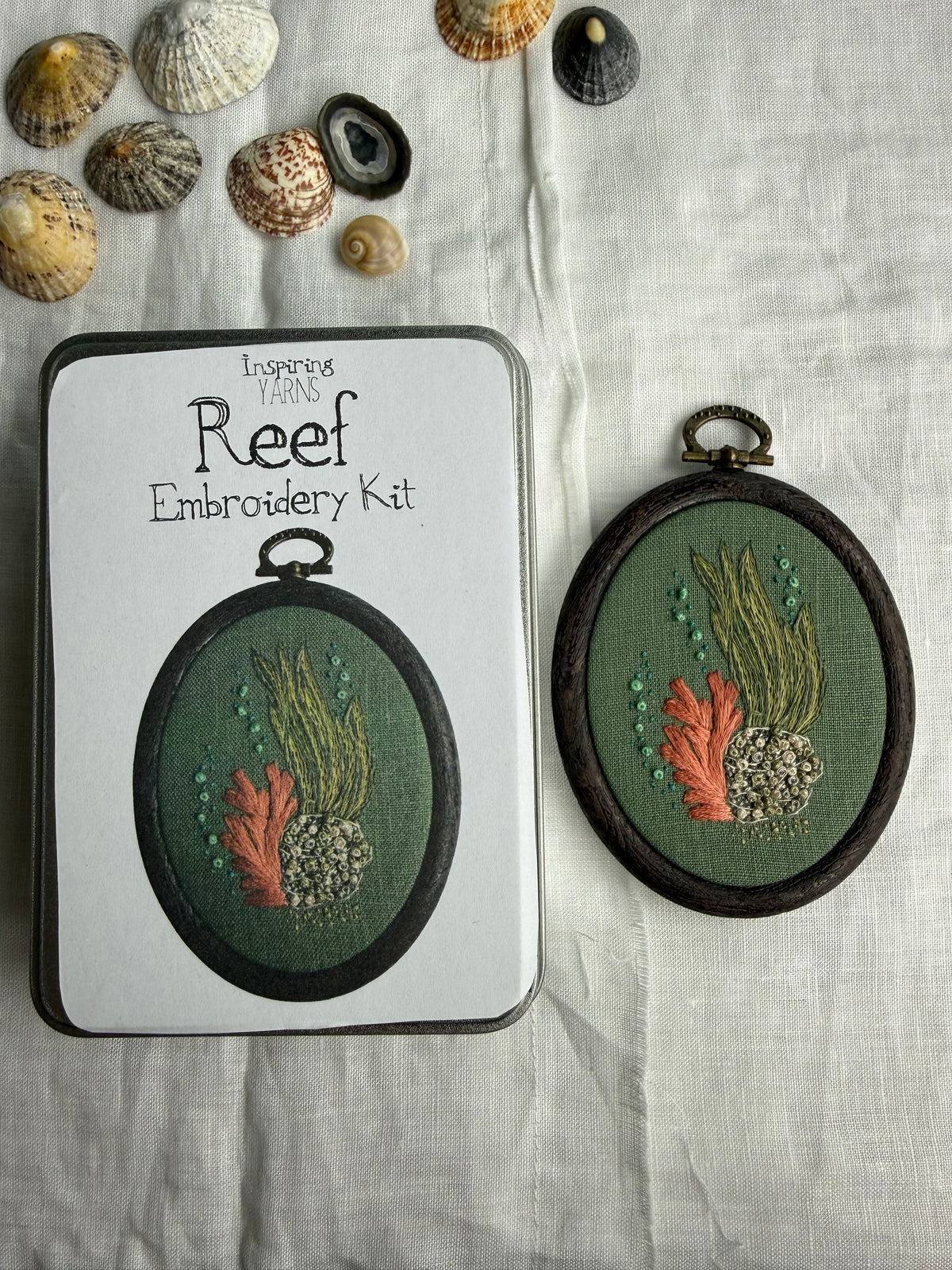 An oval embroidery kit shown with its white packaging tin and the completed embroidered piece. The design depicts coral reef motifs in green, orange, and grey tones on a dark green fabric background, framed in dark brown.