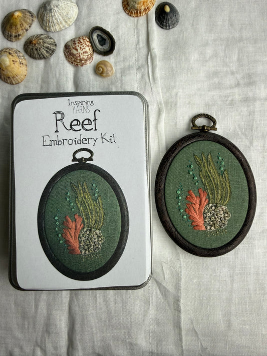 An oval embroidery kit shown with its white packaging tin and the completed embroidered piece. The design depicts coral reef motifs in green, orange, and grey tones on a dark green fabric background, framed in dark brown.