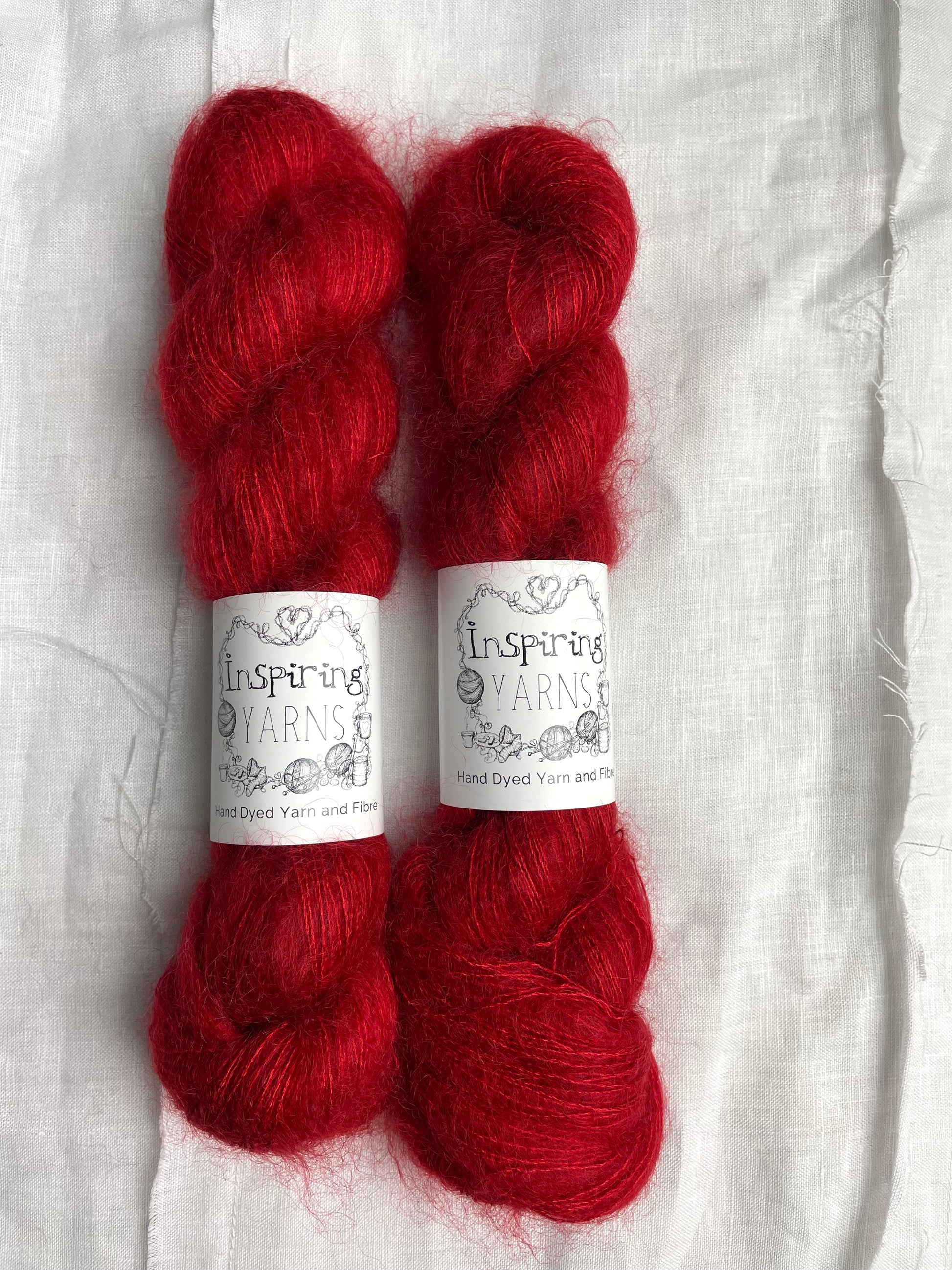 Two skeins of tonal bright red kid mohair silk lace yarn labelled Inspiring Yarns on a white linen background.