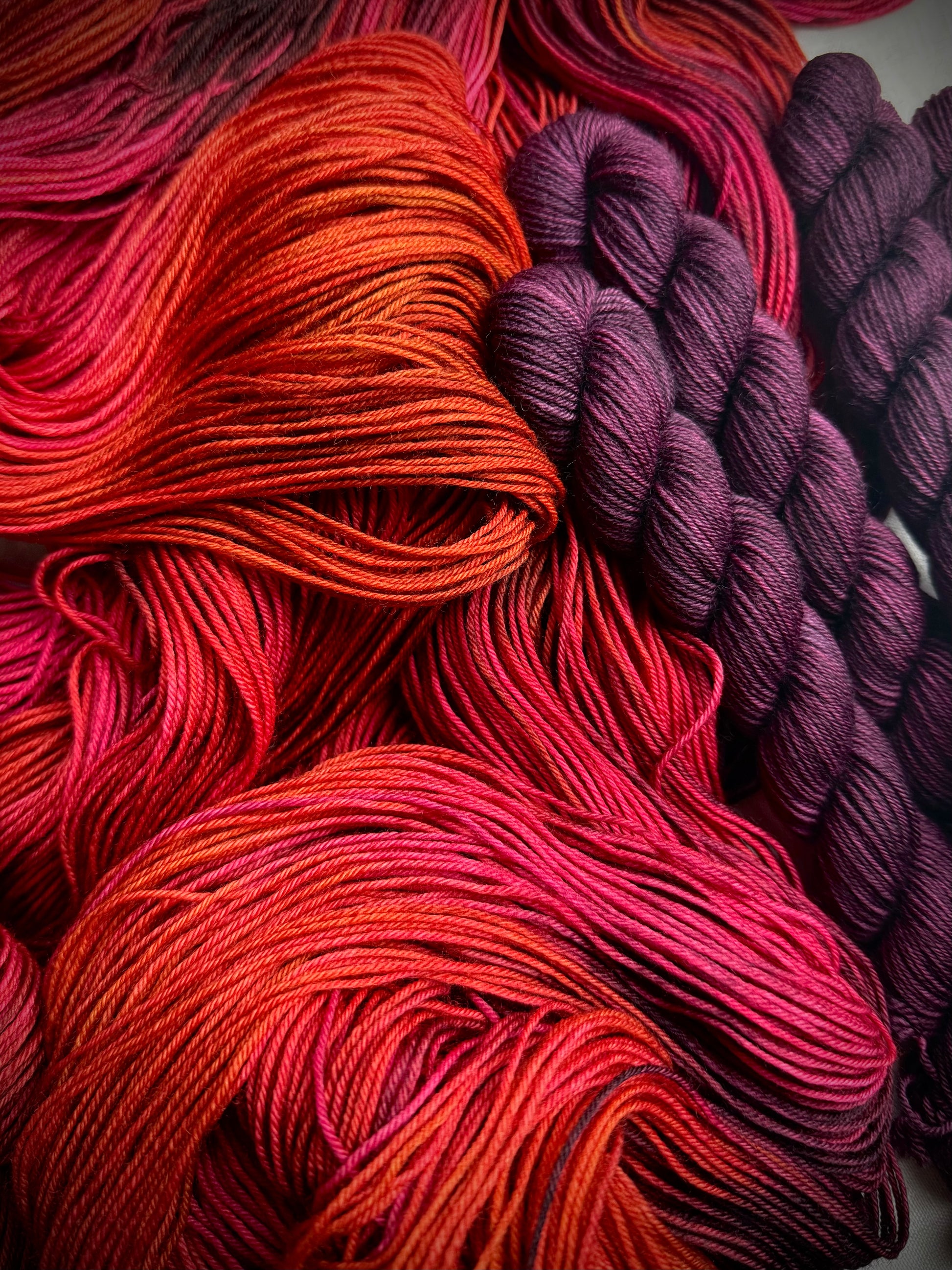 Variegated red, pink, orange and purple yarn  with a burgundy mini skein on a white fabric background