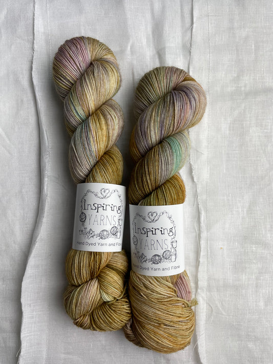 Two soft gold and mixed pastel coloured skeins of Merino Smooth Sock yarn branded  Inspiring Yarns on a white linen fabric background. 