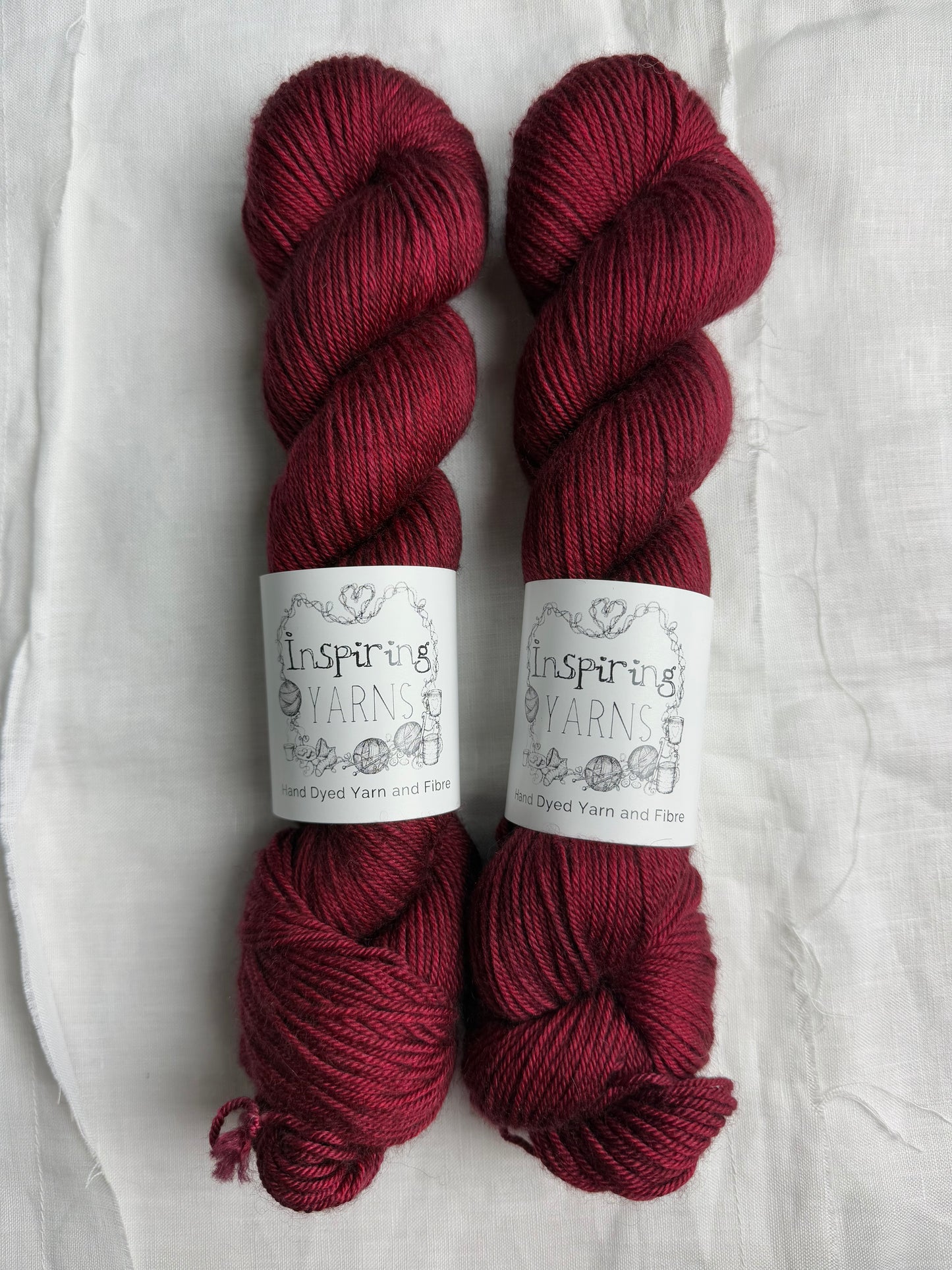 two skeins of yarn in a dark ruby red on a white background and inspiring yarns labels around them.